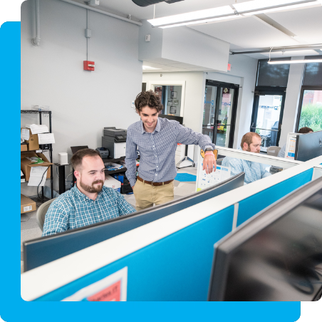 Guy standing in cubicle, while the other sits in front of computer