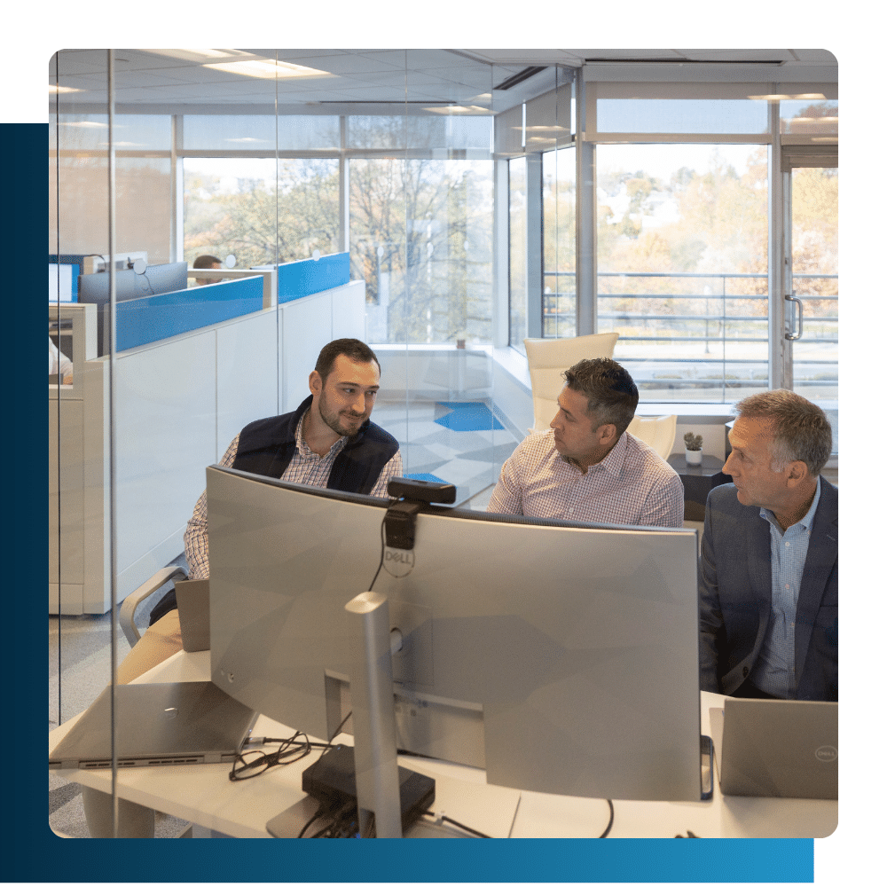 Three professionals collaborating at a computer workstation in a modern glass-walled office, discussing financial IT solutions.