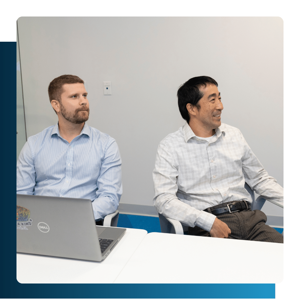 Two professionals seated at a table in a meeting room, engaged in discussion with laptops open.
