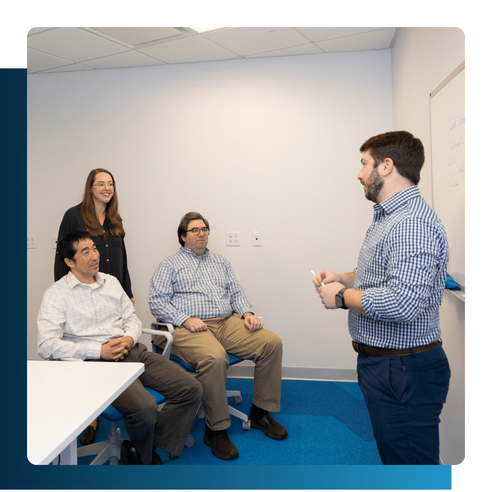 Four professionals in a meeting room engaged in a collaborative IT strategy discussion, with one team member presenting at a whiteboard.